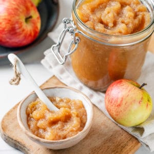 Easy Apple Jam with 2 Ingredients (homemade apple sauce) A glass jar and a white bowl of apple jam with a spoon in the bowl and apples all around.