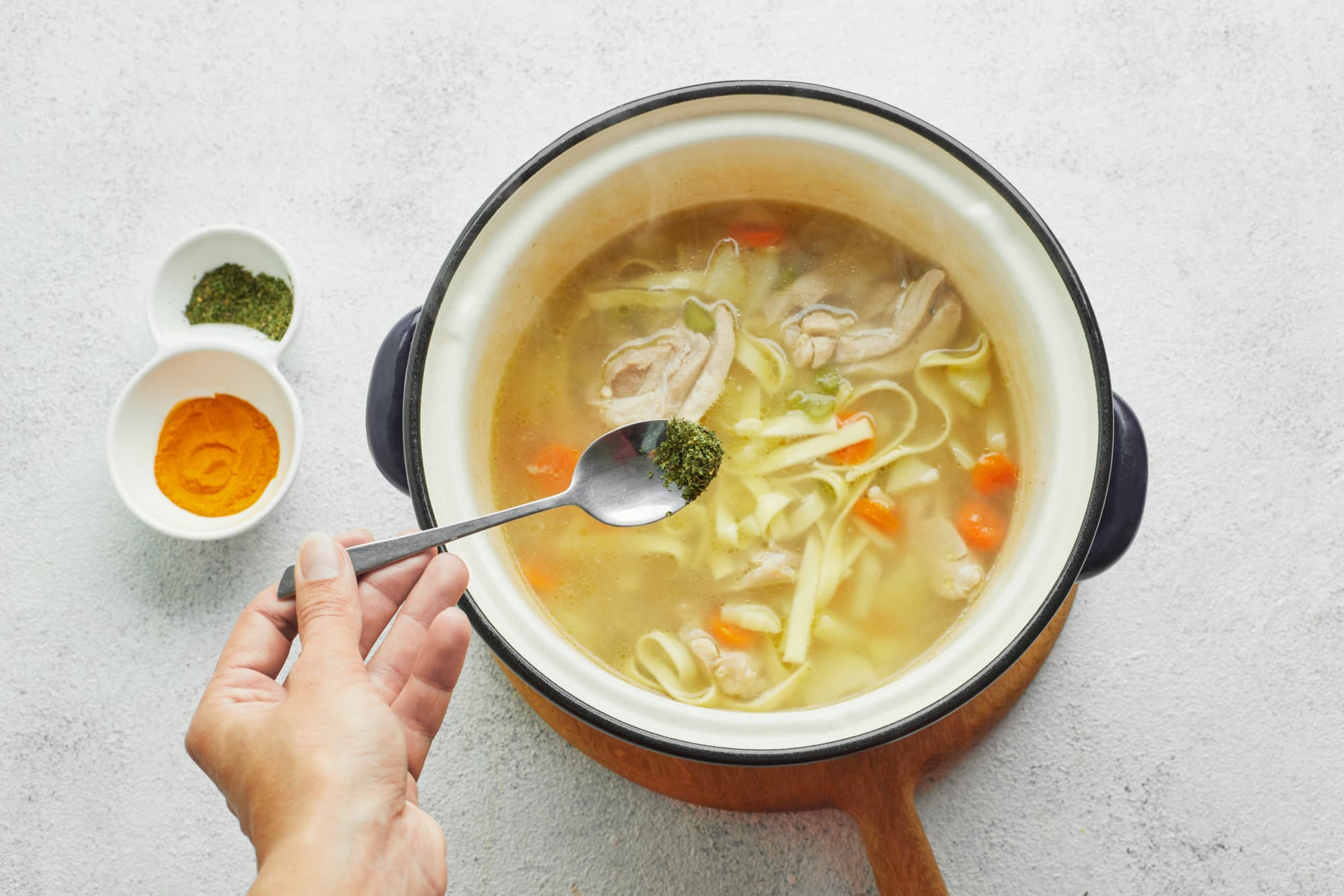 The BEST Chicken Noodle Soup Recipe (with Potatoes!) chicken-soup-in-a-pot-on-a-wooden-baord-with-spices-being-added-in-with-a-spoon-with-two-bowls-of-spices-on-the-side