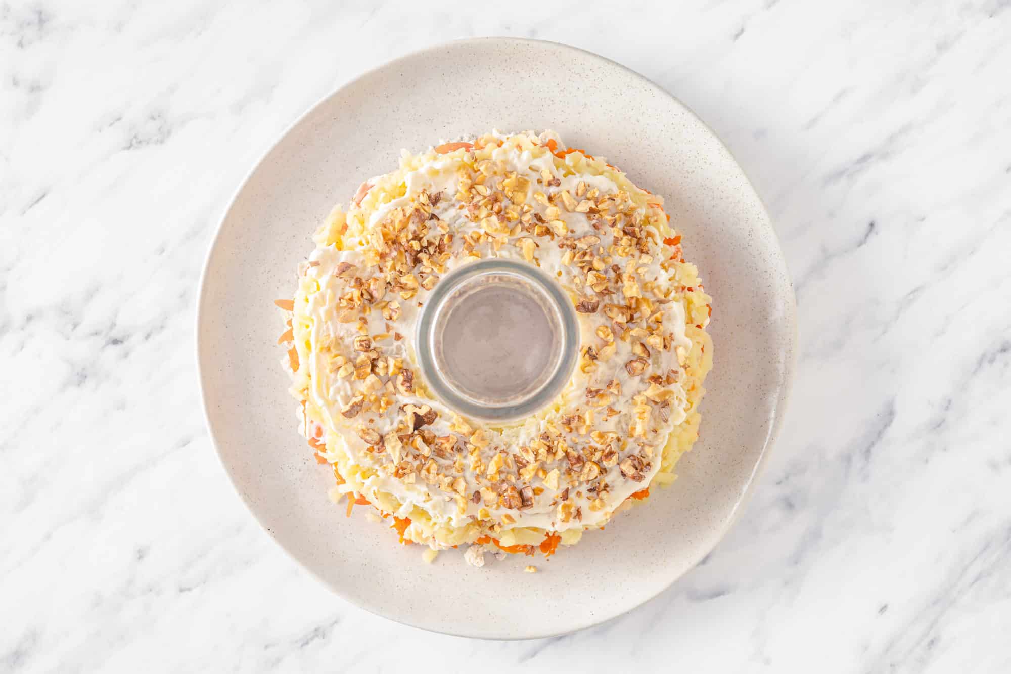 walnuts atop mayonnaise and a grated potato salad layer on a white plate with a glass cup in the center.