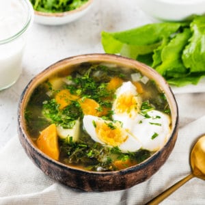 How to make Green Herb Soup with Sorrel (Shchavel Borscht) A brown bowl of sorrel soup on a white towel with a gold spoon on the side, and sorrel leaves.