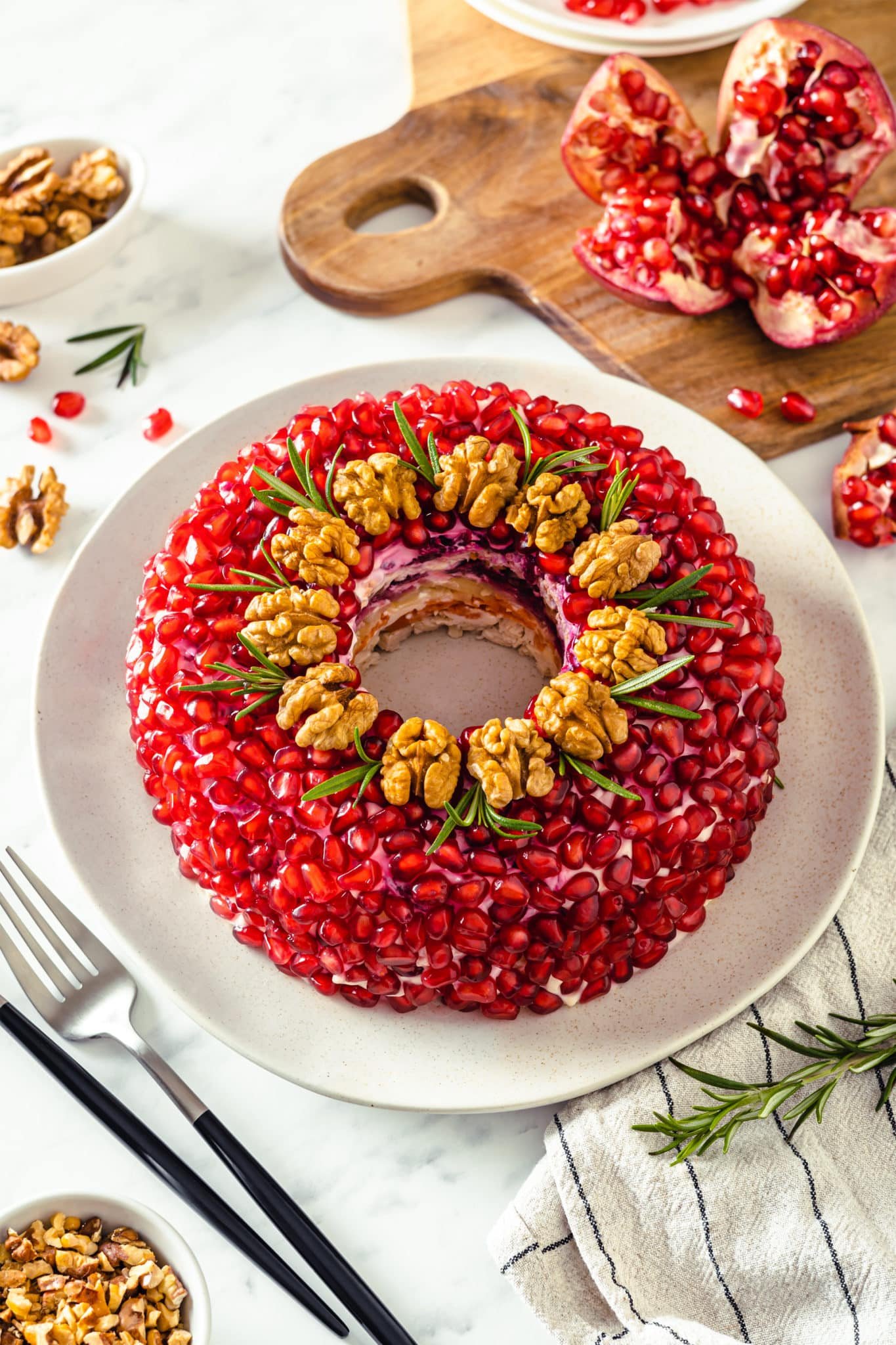 Layered Chicken Salad with Pomegranate layered chicken salad with pomegranate walnuts and rosemary on it on a white plate with a fork and a towel on the side as well as more pomegranate.