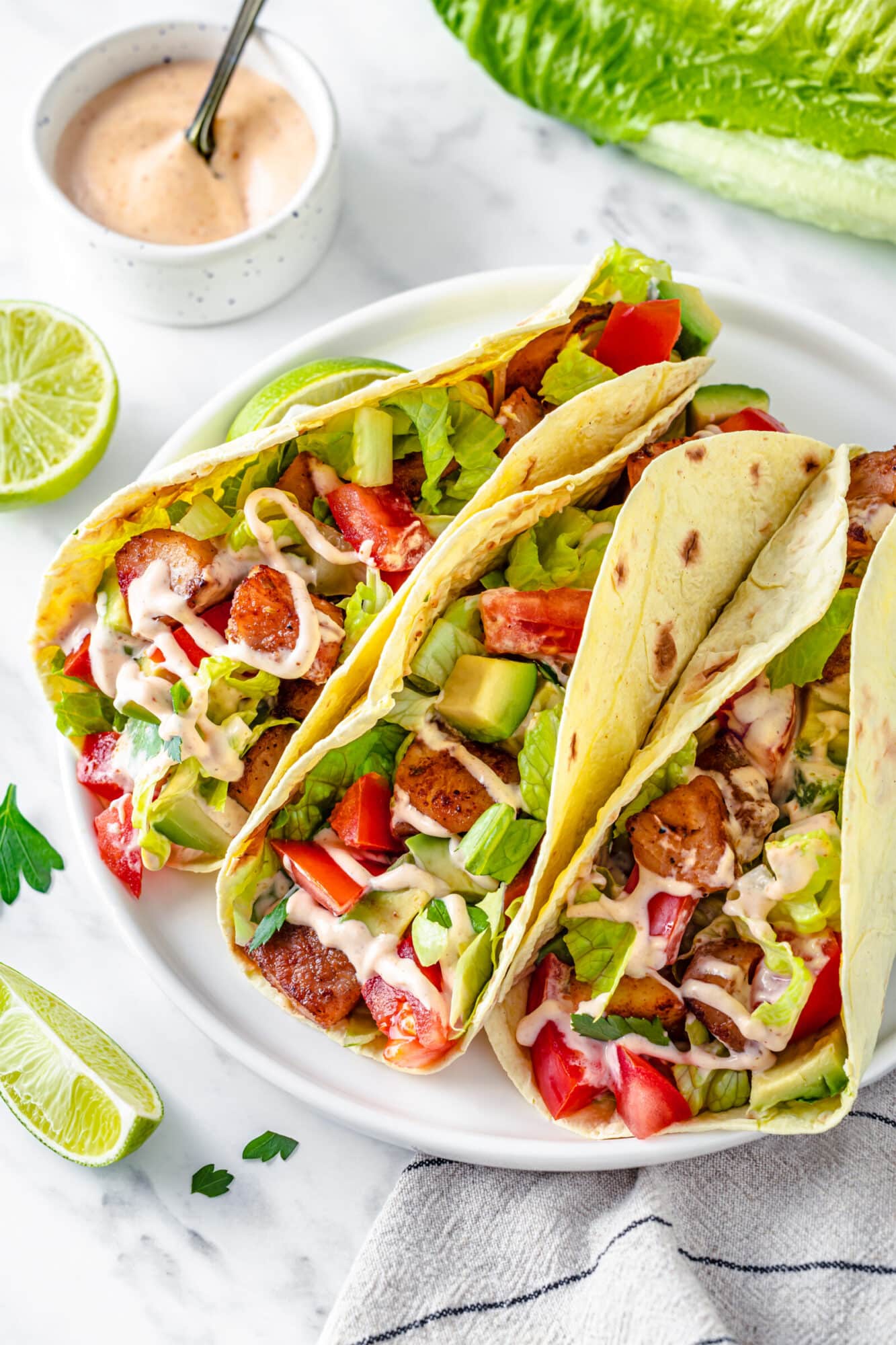 Fish Tacos with an Easy Spicy Dipping Sauce Soft taco shells filled with fish, chopped veggies, and drizzled with spicy fish sauce with lime wedges on the side for serving.