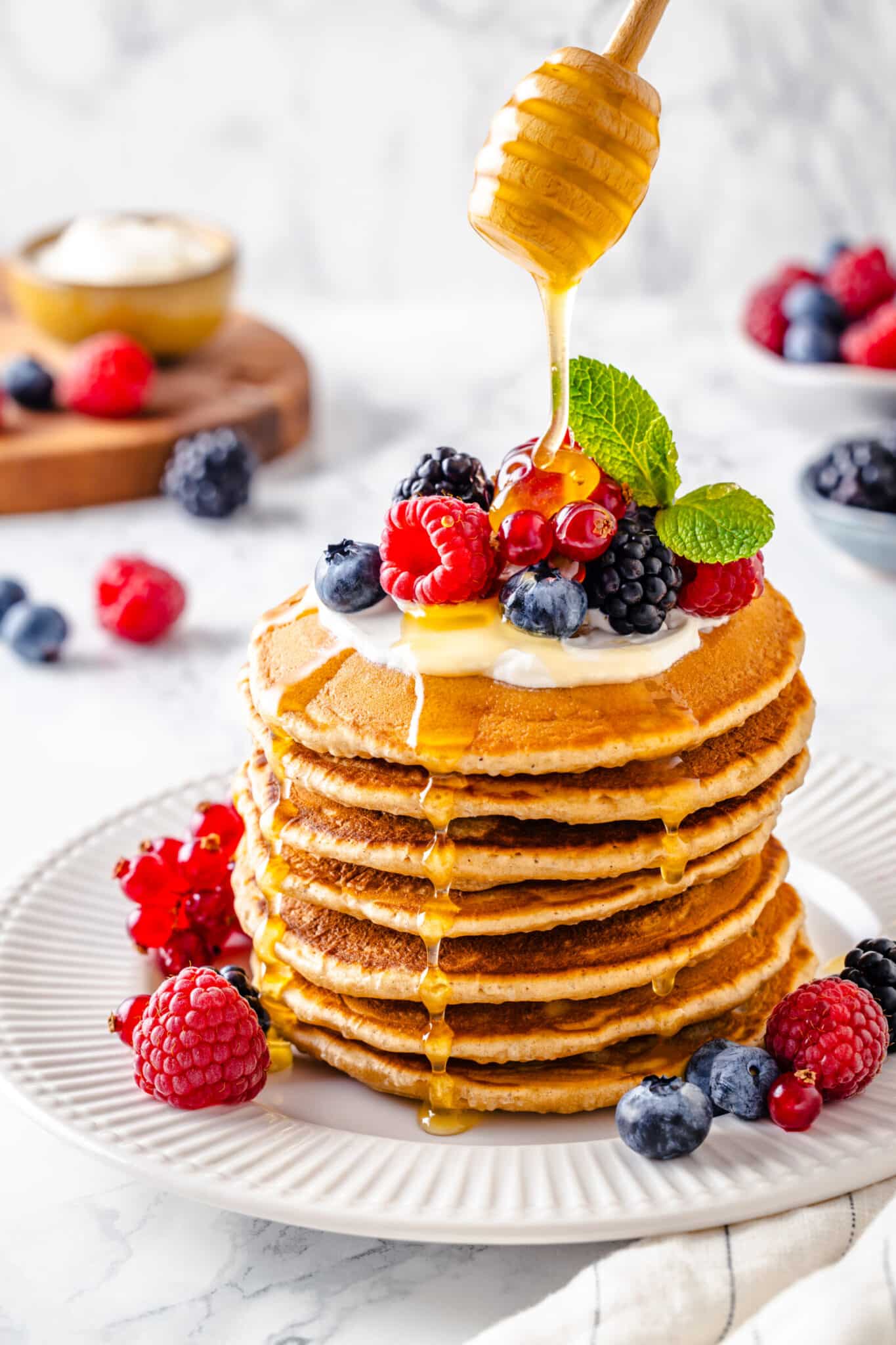The BEST Fluffy Pancake Recipe a large stack of thick pancaked with cream and berries on top and around it, with honey being drizzled over it